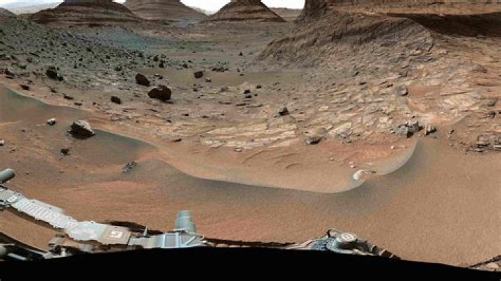 The Curiosity rover took this photo in 2012, showcasing its own three left wheels. In the distance you can see the lower slope of Mount Sharp